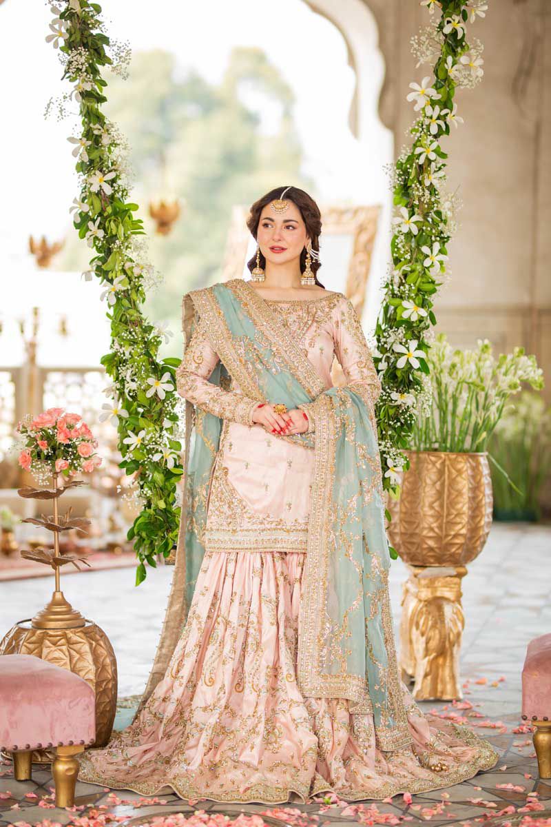 Model wearing Hania Embroidered Net Gharara in baby pink with intricate stitching and embellishments in a scenic outdoor setting.