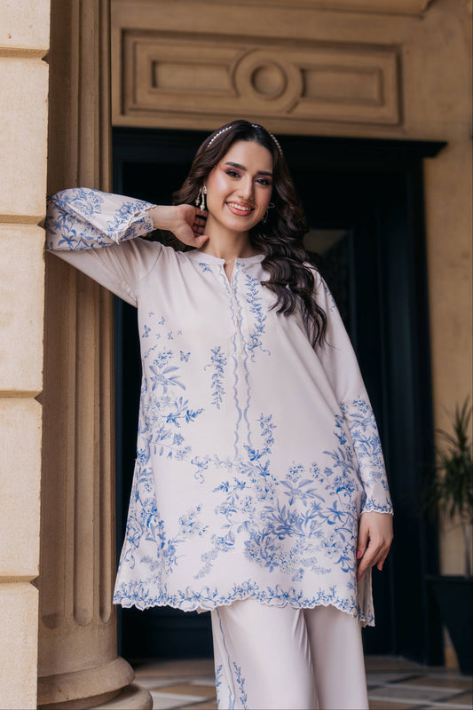 A woman models a light beige or off-white three-piece kurta set with blue floral embroidery, standing in front of a classical stone doorway.
