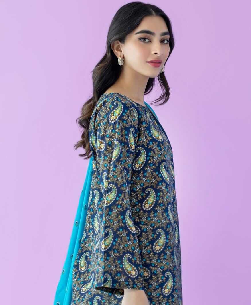 A woman modeling a dark blue printed kurta with intricate paisley and floral patterns in green, gold, and white, paired with a bright turquoise dupatta, set against a solid lavender background.