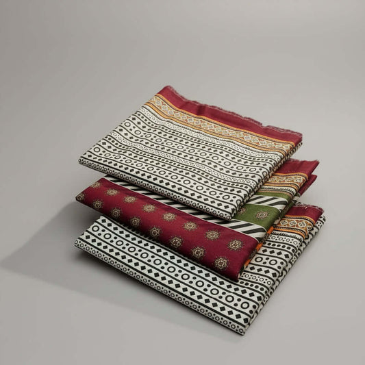 Three folded patterned fabric pieces with maroon, black, white, and green geometric and floral designs, arranged on a white faux-fur surface. A small white tag with a pink logo is attached to the lower fabric.