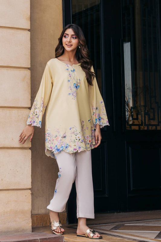 A woman models a beige tunic with blue and pink floral embroidery, paired with light beige pants and metallic sandals, standing in front of a beige stone wall and a black door.