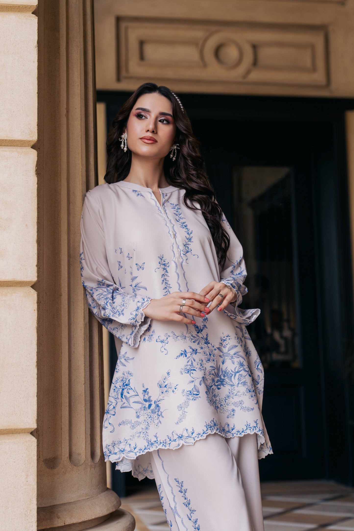 Woman in a light-colored traditional outfit with floral patterns standing in front of a building entrance.