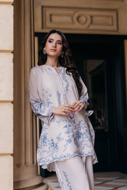 Woman in a light-colored traditional outfit with floral patterns standing in front of a building entrance.