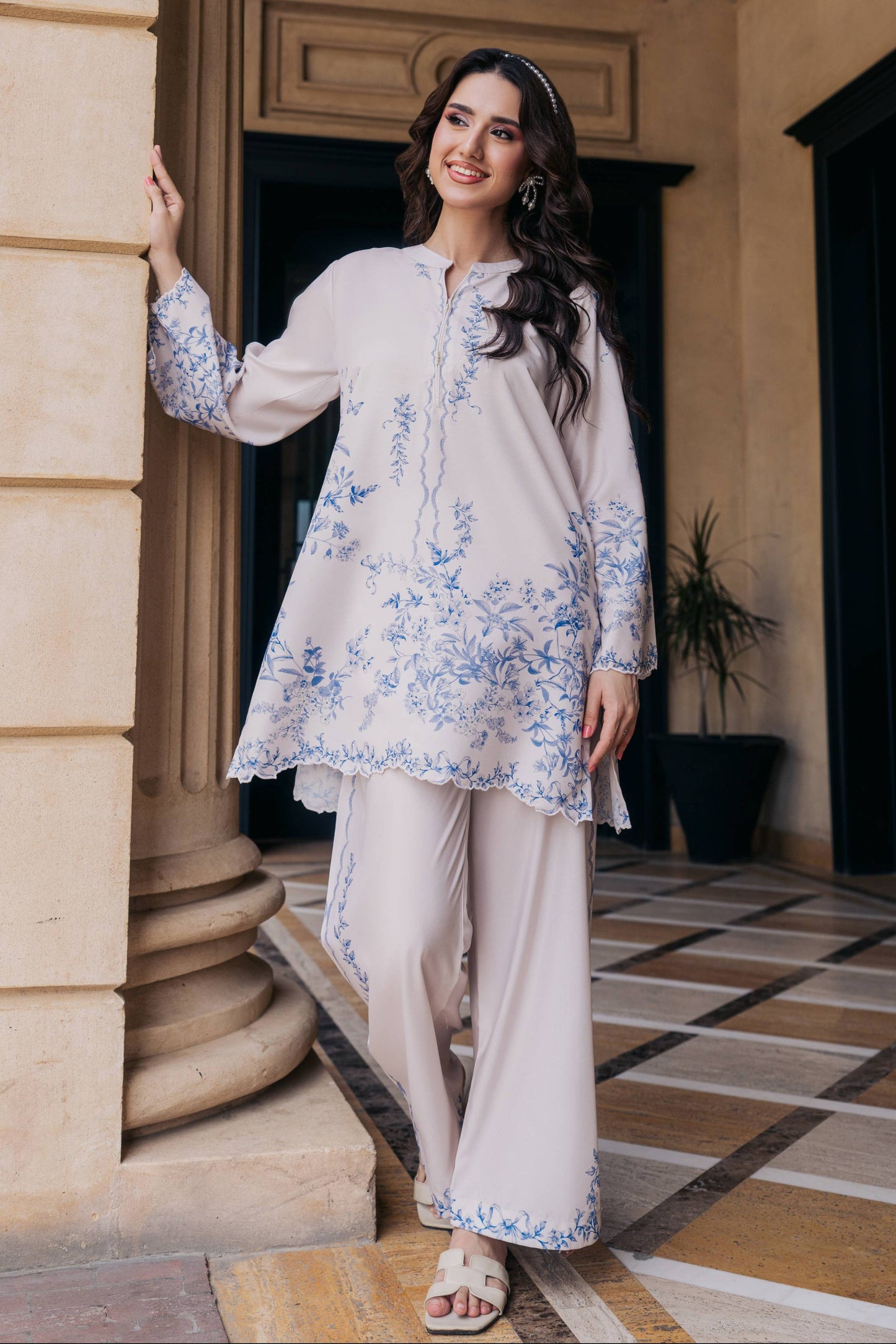 A woman models a traditional three-piece kurta set in a beige color with blue floral embroidery, standing in front of a beige stone column.