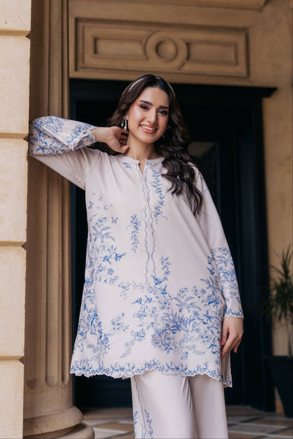 A woman models a light beige or off-white three-piece kurta set with blue floral embroidery, standing in front of a classical stone doorway.