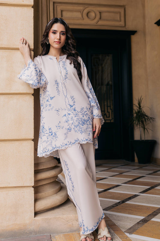 A woman models a beige two-piece kurta and trouser set with blue floral embroidery, standing in a warm-toned stone corridor.