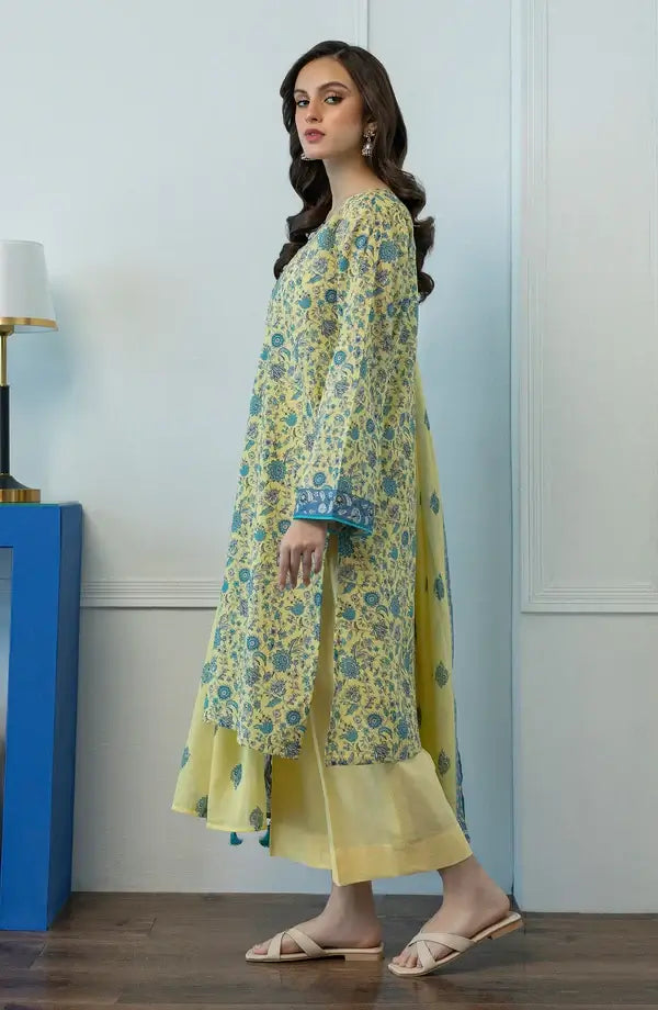 A woman modeling a yellow and blue floral printed kurta set with matching pants in an indoor setting with a light blue paneled wall and a blue side table.