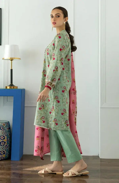 A woman models a light green three-piece salwar kameez suit with floral embroidery, matching green trousers, and a pink dupatta with gold floral patterns, standing in a modern room with a blue side table and lamp.