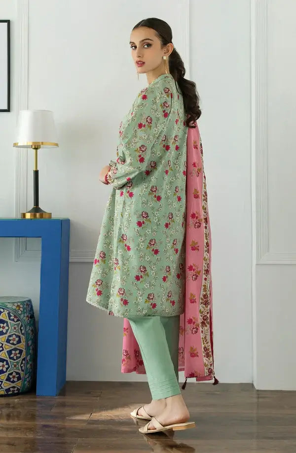 A woman models a light green kurta with red and gold floral embroidery, paired with mint green pants and a pink dupatta with gold borders, standing in a room with a blue side table and lamp.