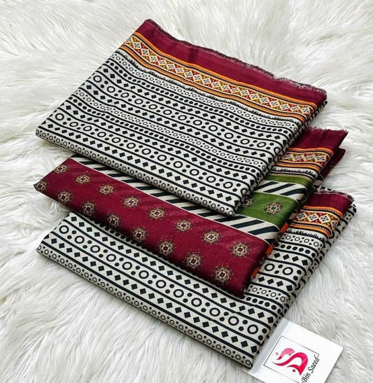 Three folded patterned fabric pieces with maroon, black, white, and green geometric and floral designs, arranged on a white faux-fur surface. A small white tag with a pink logo is attached to the lower fabric.