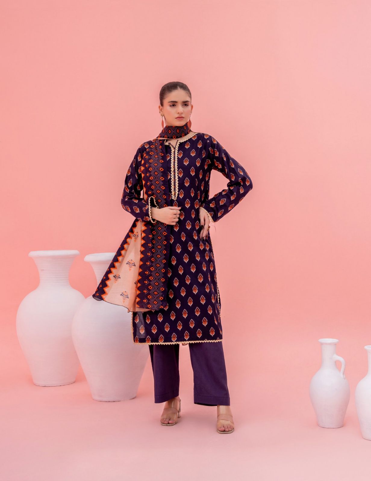 Woman in a patterned outfit standing against a pink background with white vases.